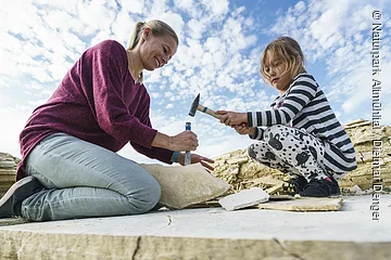 Frau und Mädchen schlagen mit Hammer und Meißel auf Steinplatte bei Ausgrabung im Freien ein.