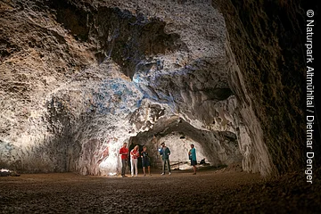 Fünf Personen stehen in einer großen, beleuchteten Höhle mit unregelmäßigen Felswänden und Boden aus Erde.
