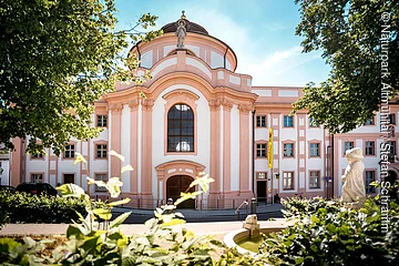 Barockgebäude mit großer Fensterfront, Statue auf dem Dach und Springbrunnen im Vordergrund bei Sonnenschein