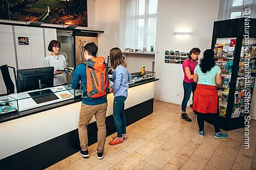 Innenraum einer Touristeninformation mit vier Personen, zwei am Tresen und zwei vor einem Prospektständer.