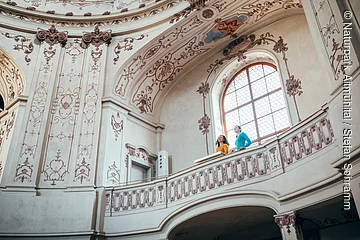 Zwei Personen stehen auf einem verzierten Balkon in einem historischen, mit Fresken geschmückten Innenraum.