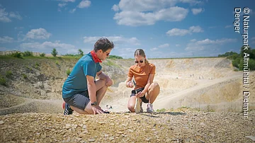 Zwei Personen knien auf einem steinigen Gelände und untersuchen den Boden.