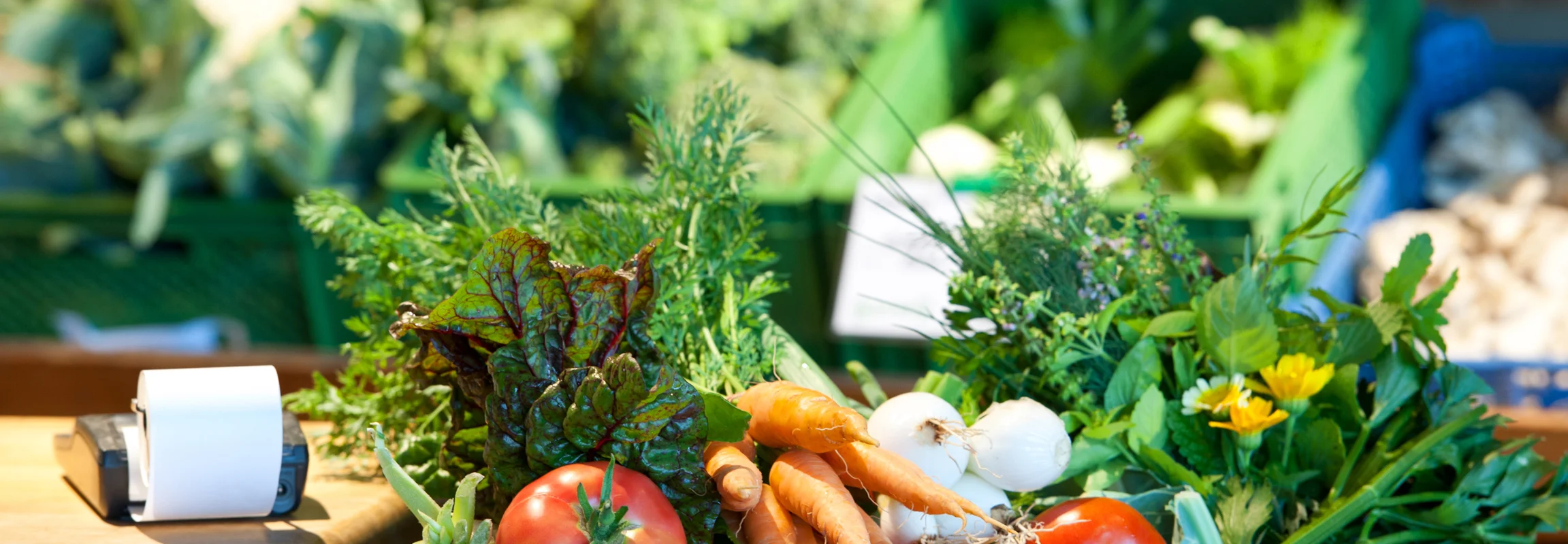 Holzkiste mit frischem Gemüse wie Tomaten, Karotten, Zwiebeln und Kräutern auf einem Marktstand.