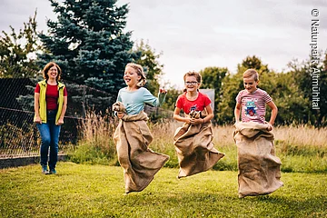 Drei Kinder springen in Jutesäcken auf einer Wiese, eine Frau beobachtet sie lachend im Hintergrund.