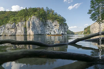 Fluss mit Felsen und Bäumen, Boot mit zwei Personen, Äste im Vordergrund im Wasser sichtbar