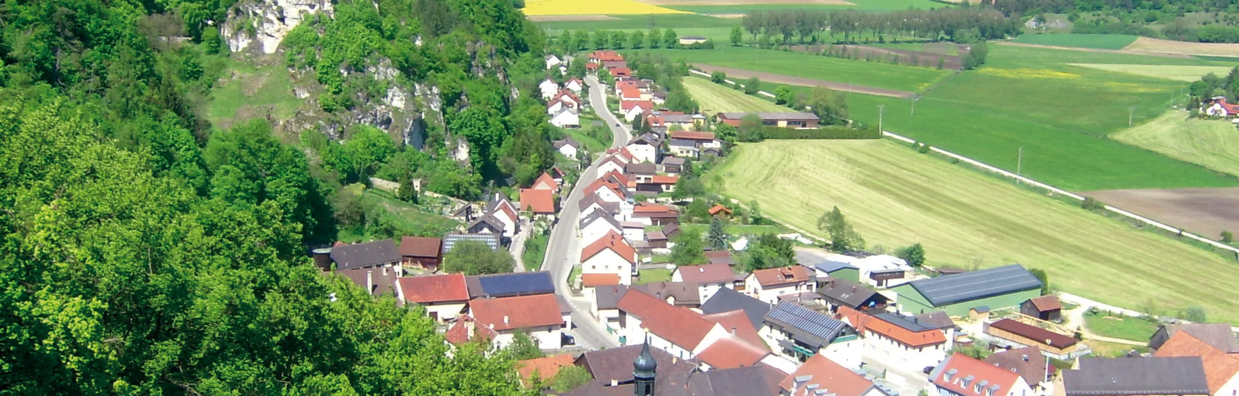 Blick auf ein Dorf mit Kirche und Burgruine auf bewaldetem Hügel, umgeben von Feldern und Wiesen.