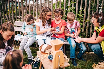 Kinder und Erwachsene sitzen auf einer Bank im Garten und bearbeiten Getreide mit einer Mühle aus Holz.