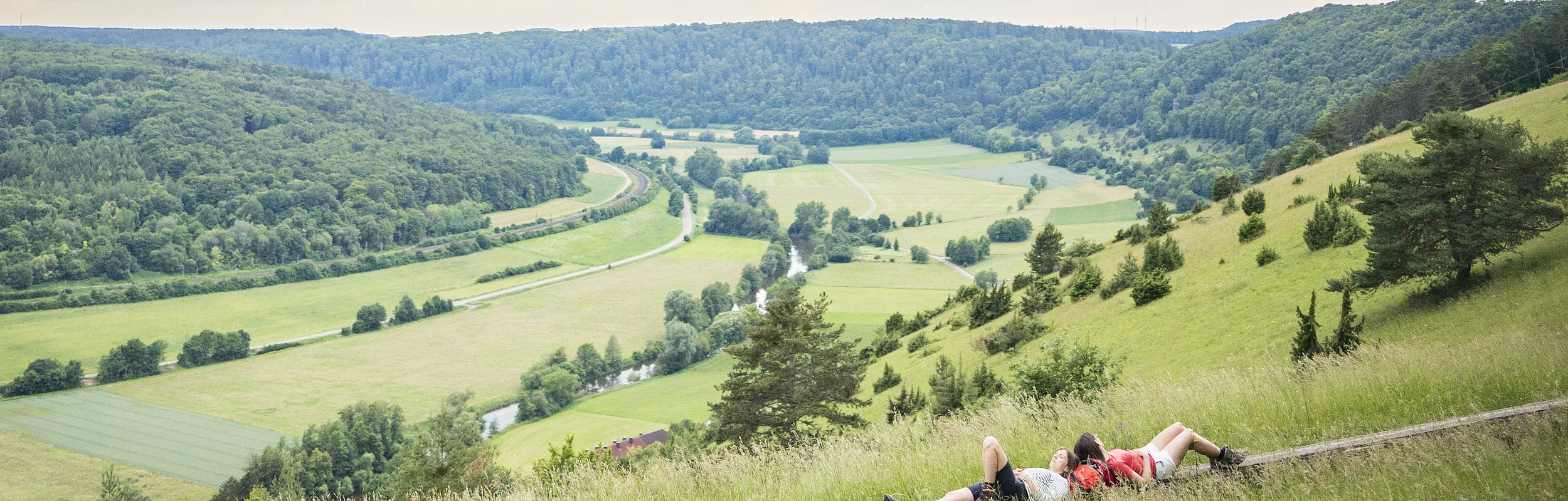Zwei Wanderinnen liegen auf einer langen Holzbank und schauen ins weitläufige grüne Altmühltal.