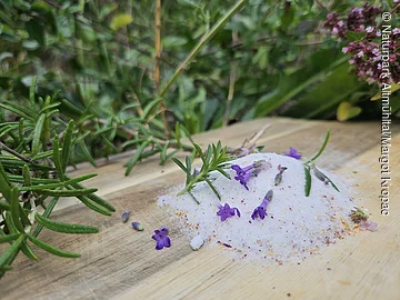 Haufen Salz mit kleinen lila Blüten und grünen Kräutern auf Holzbrett, unscharfer Hintergrund mit Pflanzen.