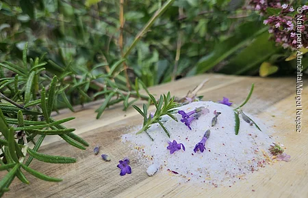 Haufen Salz mit kleinen lila Blüten und grünen Kräutern auf Holzbrett, unscharfer Hintergrund mit Pflanzen.