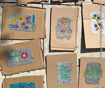 Mehrere Hand geschöpfte Papierstücke mit Blumen- und Schmetterlingsmotiven liegen auf braunen Kartonstücken auf einem Steinboden.