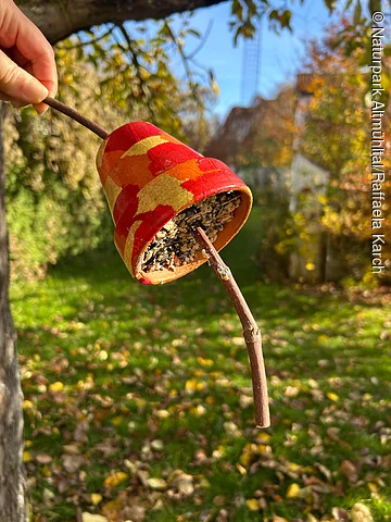 Hand hält bemalten Blumentopf mit Vogelfutter an einem Ast vor unscharfem Gartenhintergrund im Herbst