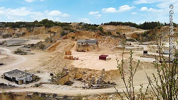 Steinbruch mit Sand- und Felsformationen, Gebäuderuinen und Paletten, umgeben von Vegetation.