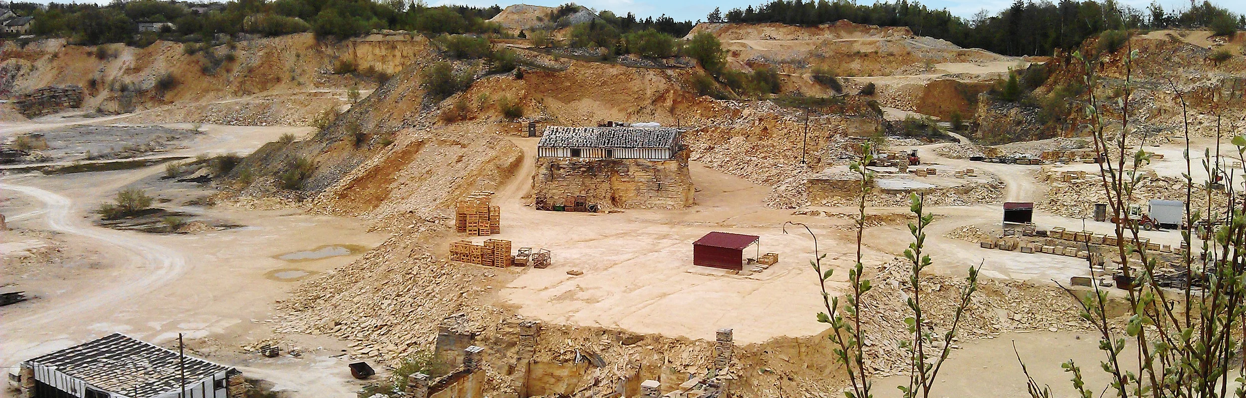 Steinbruch mit Sand- und Felsformationen, Gebäuderuinen und Paletten, umgeben von Vegetation.