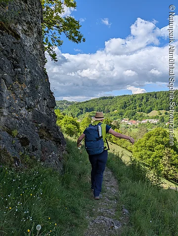 Wanderer mit Rucksack und Hut auf schmalem Pfad neben Felswand, grüne Hügel und Dorf im Hintergrund.