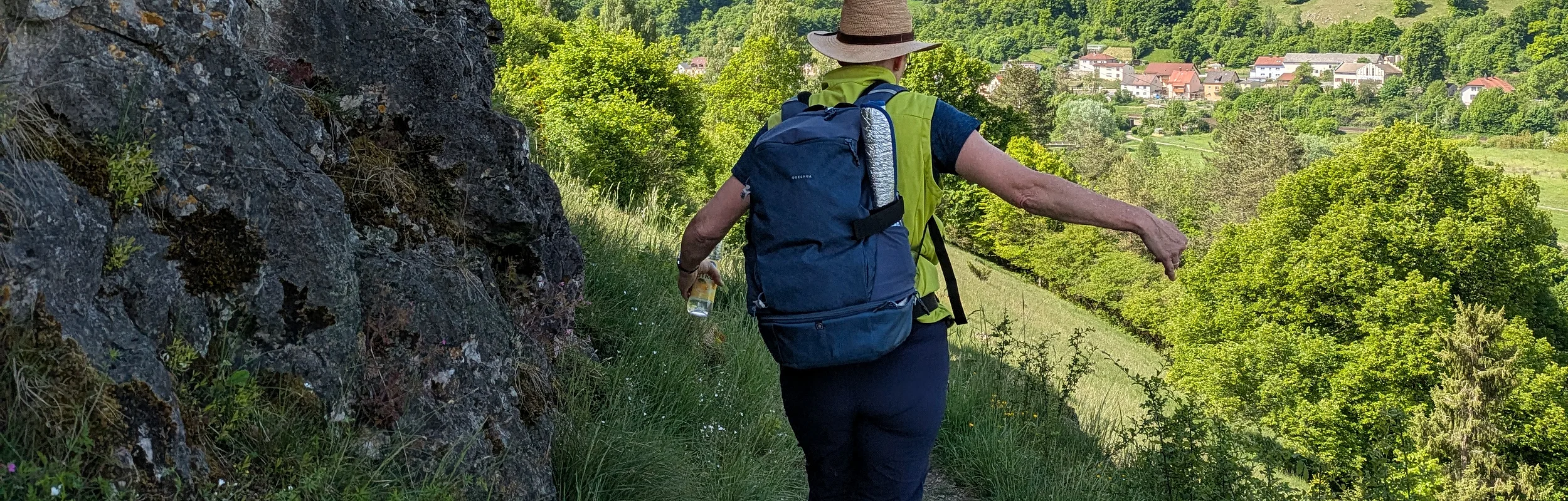 Wanderer mit Rucksack und Hut auf schmalem Pfad neben Felswand, grüne Hügel und Dorf im Hintergrund.