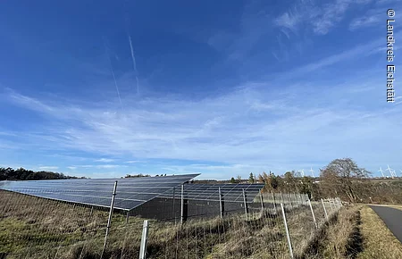 Solarpaneele auf einer Wiese hinter einem Metallzaun unter blauem Himmel mit Windrädern im Hintergrund