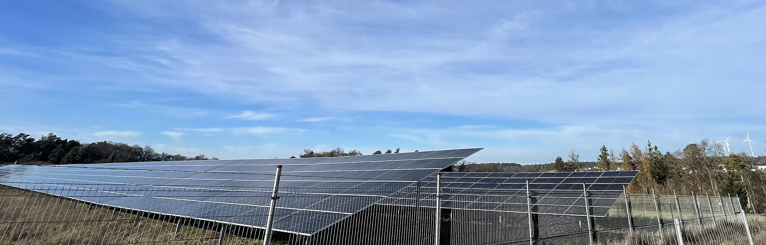 Solarpaneele auf einer Wiese hinter einem Metallzaun unter blauem Himmel mit Windrädern im Hintergrund