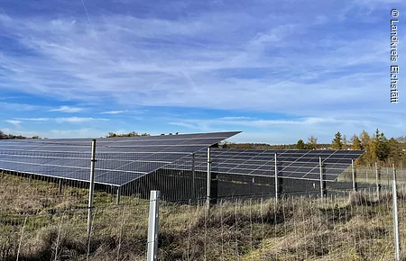 Solaranlage mit mehreren Solarpanelen hinter einem Metallzaun auf einer Wiese unter blauem Himmel.