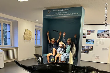 Drei Kinder sitzen vor einer blauen Museumsvitrine mit Text, davor ein großes Modell eines urzeitlichen Meeresreptils.