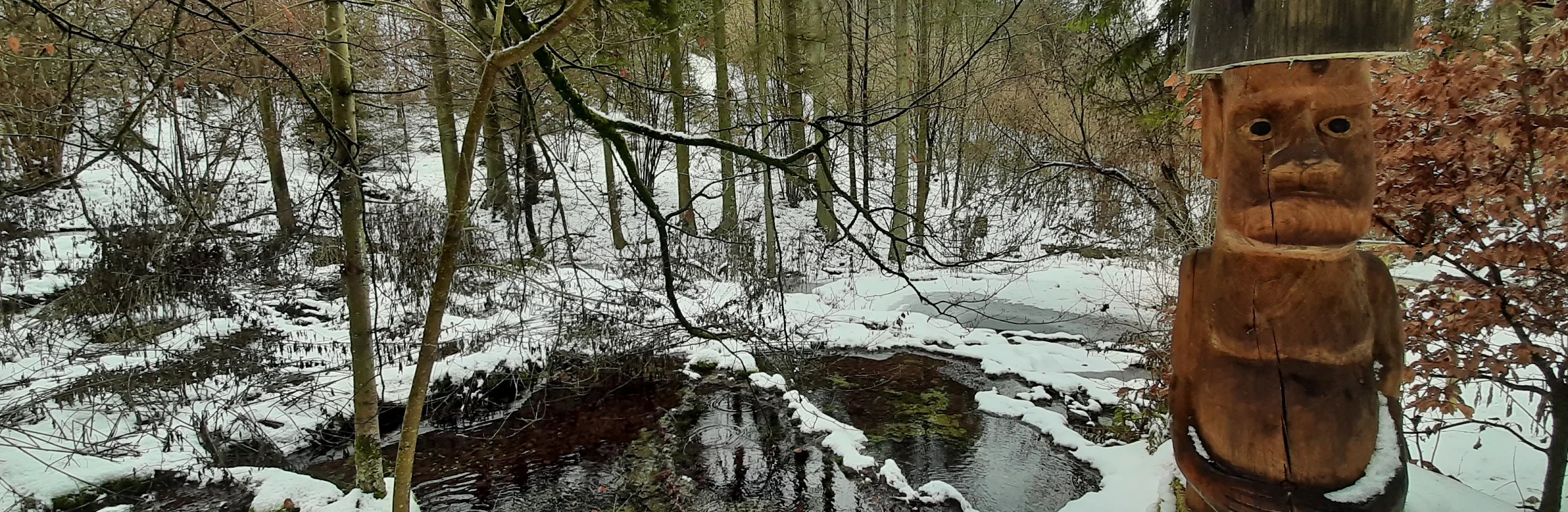 Holzfigur mit Schneehut neben einem kleinen Bach im verschneiten Wald im Winter.