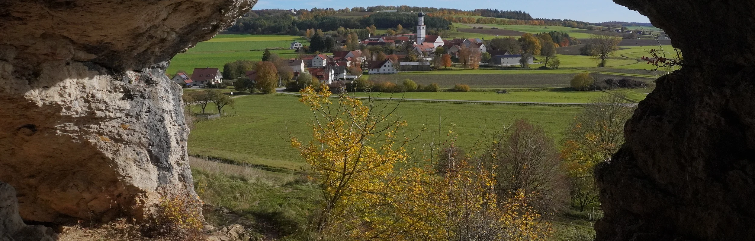 Blick aus einer Höhle auf ein Dorf mit Kirche, Feldern und Bäumen im Herbst bei klarem Himmel.
