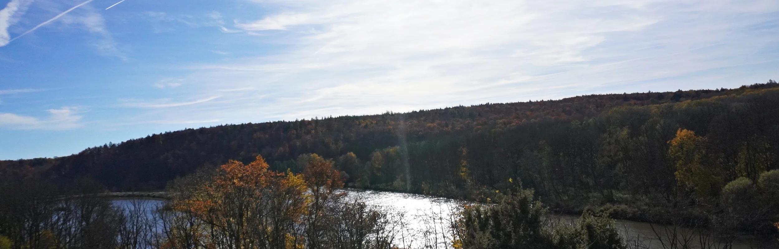 Fluss mit Bäumen am Ufer und bewaldetem Hügel unter blauem Himmel mit Sonne und Wolken.