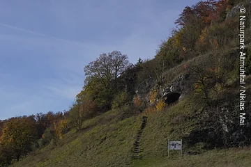Grasbewachsener Hang mit Treppe, Bäumen und einer Felsöffnung unter blauem Himmel.