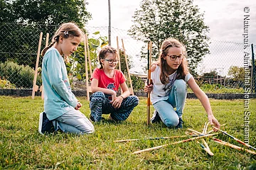Drei Mädchen knien auf Gras und spielen mit langen Holzstäben im Garten vor einem Zaun.