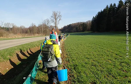 Kinder mit Rucksäcken und Eimern gehen auf einem Feldweg neben einer Straße und Wiese entlang.