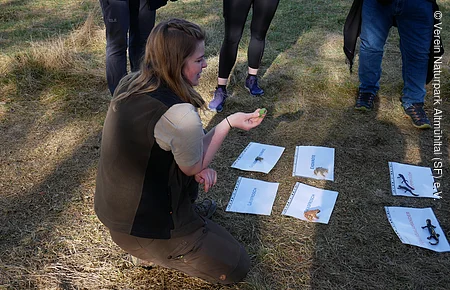 Frau kniet auf Wiese, hält Frosch, umgeben von drei stehenden Personen und Tierbildern auf Boden.