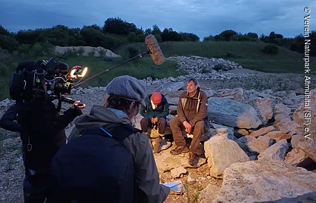Filmteam mit Kamera und Mikrofon filmt zwei Personen, die auf Felsen in einer Landschaft sitzen.
