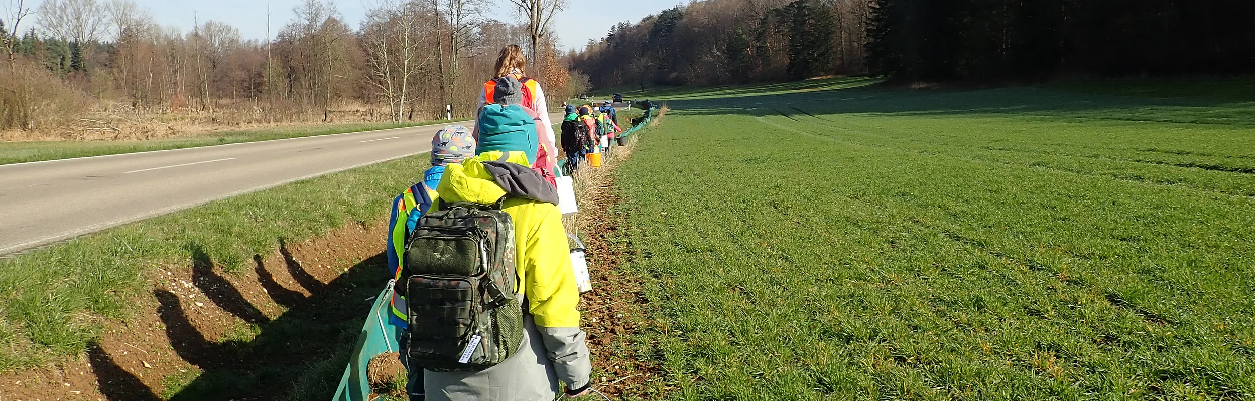 Kinder mit Rucksäcken und Eimern gehen auf einem Feldweg neben einer Straße und Wiese entlang.