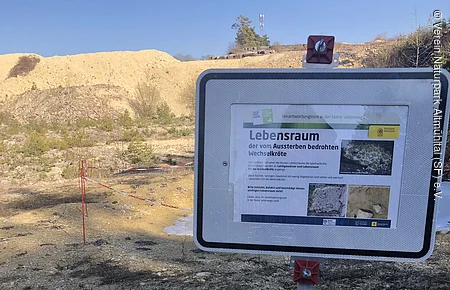 Hinweisschild zu Lebensraum der vom Aussterben bedrohten Wechselkröte in kiesigem Gelände mit Absperrung.