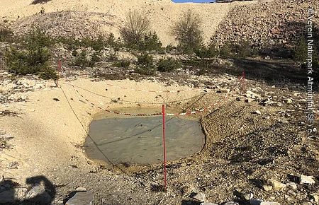 Wasserbecken auf kiesigem Boden in Steinbruch, abgesperrt mit rot-weißen Ketten an roten Pfosten.