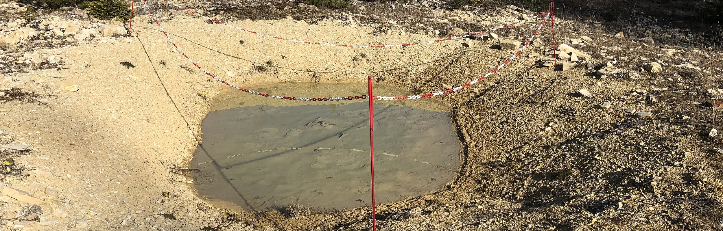 Wasserbecken auf kiesigem Boden in Steinbruch, abgesperrt mit rot-weißen Ketten an roten Pfosten.