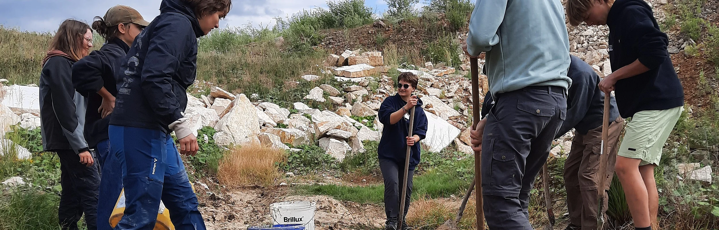 Gruppe von sieben Personen mit Schaufeln arbeitet auf einem felsigen, bewachsenen Gelände unter bewölktem Himmel.