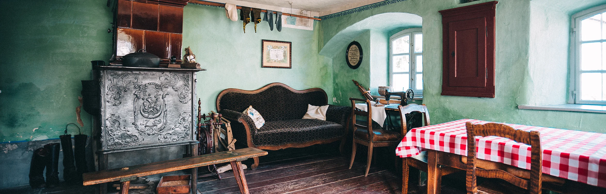Rustikaler Raum mit grün gestrichenen Wänden, einem Ofen, einer Couch, einem Holztisch mit rot-weiß kariertem Tuch und Stühlen.