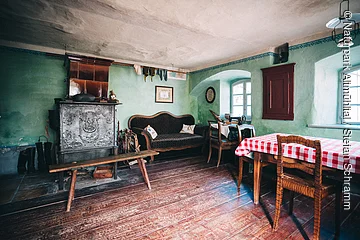 Jura-Bauernhof-Museum Hofstetten Rustikaler Raum mit grün gestrichenen Wänden, einem Ofen, einer Couch, einem Holztisch mit rot-weiß kariertem Tuch und Stühlen.
