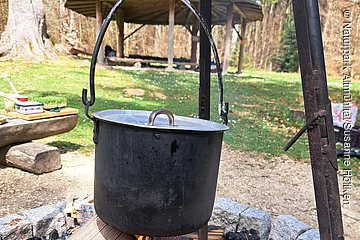 Schwarzer Topf hängt über offenem Lagerfeuer im Wald, im Hintergrund Holzhütte und Wiese.