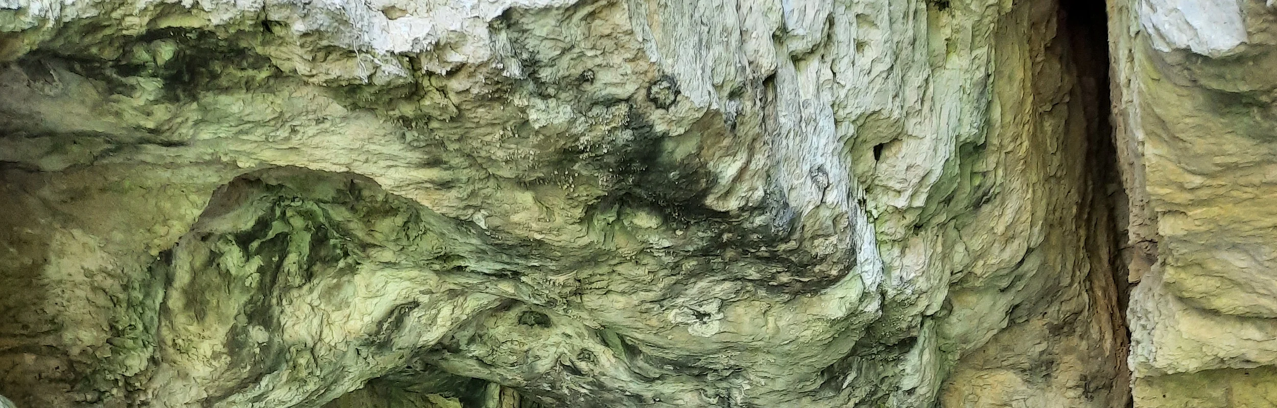Eingang einer natürlichen Höhle mit unebenem Boden und felsigen Wänden in verschiedenen Grautönen.