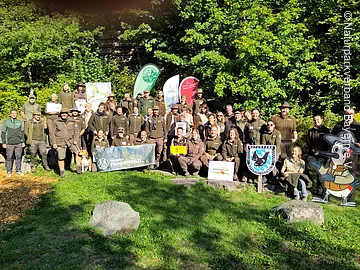 Gruppenfoto von etwa 40 Personen in Outdoor-Bekleidung vor Bäumen mit Bannern und Schildern von Naturparks.
